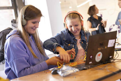 Zwei lachende Mädchen sitzen mit Kopfhörern vor einem Laptop und stecken Kabel zusammen.