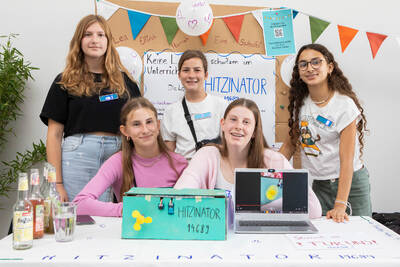Eine Gruppe Mädchen sitzt und steht hinter einem Tisch. Vor ihnen steht ein selbstgebauter Prototyp aus Pappe, der 